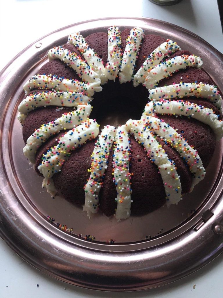 A red circular cake with rainbow sprinkles and frosting stripes.